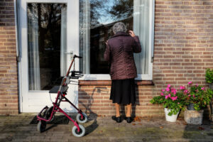 Oude dame met rollator kijkt van buiten door een raam een woning in